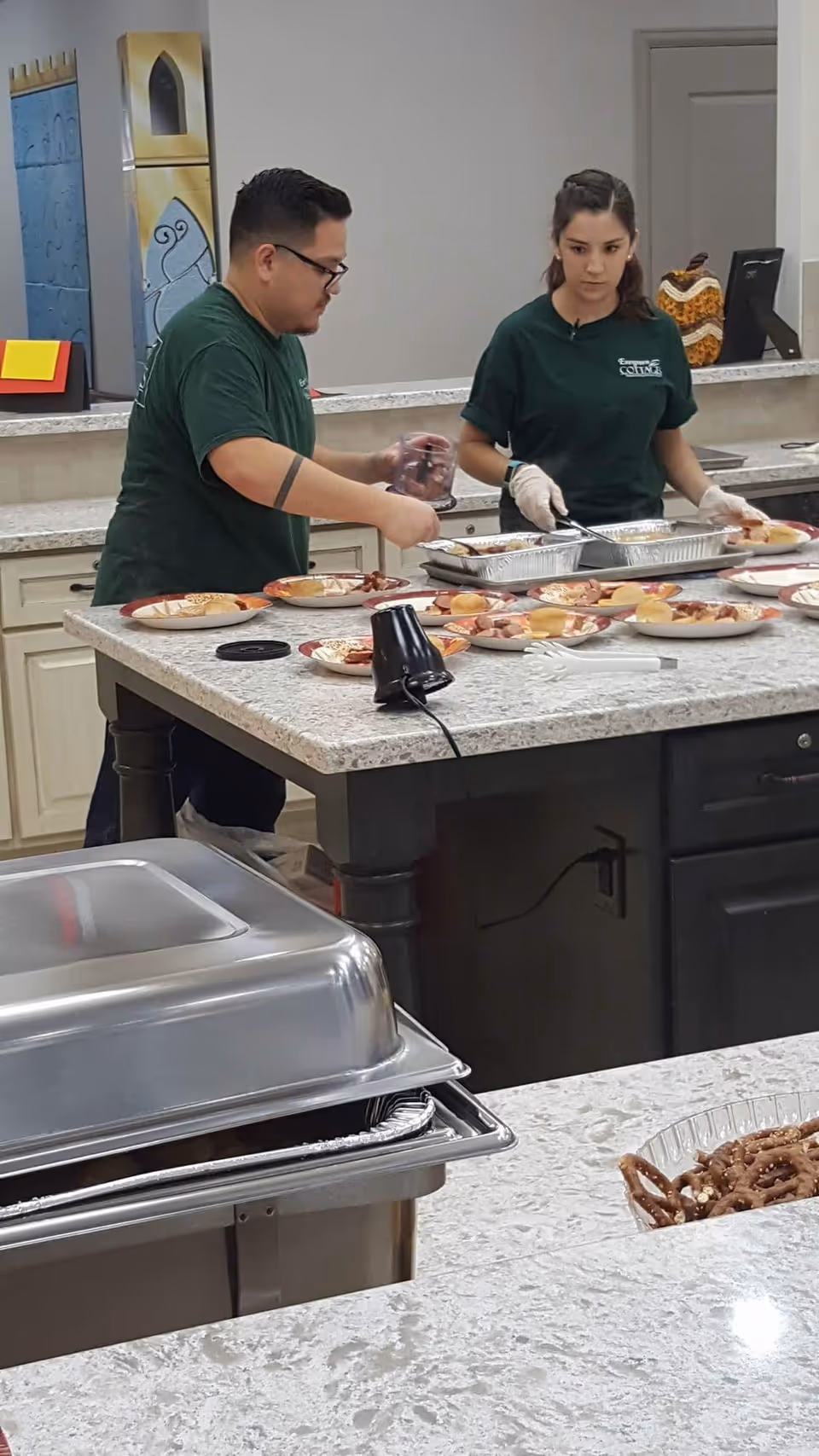 Two staff members wearing green Evergreen Cottages Magnolia shirts are preparing plates of food on a kitchen island counter. The counter has several plates with sandwiches and other food items, and there is a tray with a metal lid in the foreground.