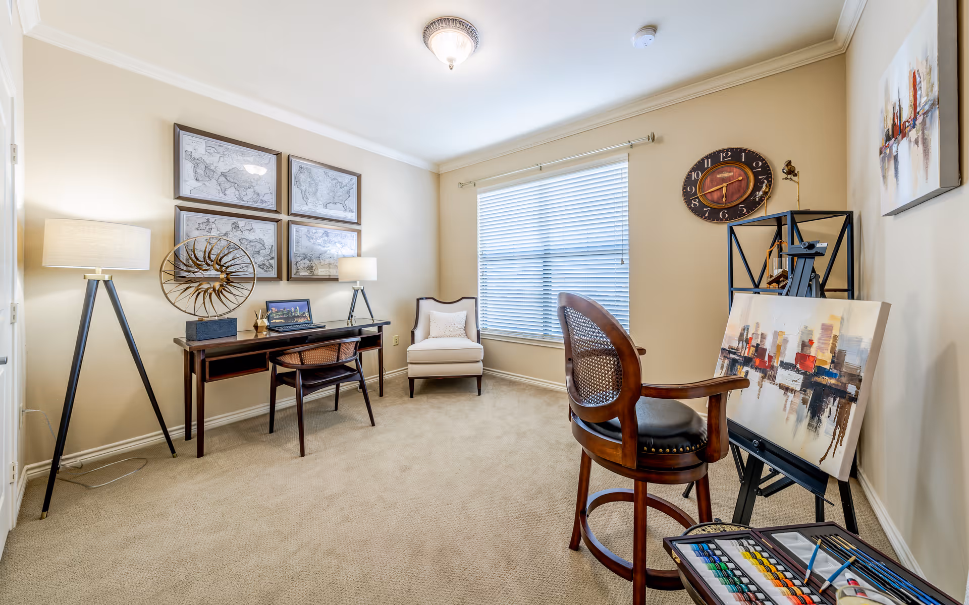 A well-lit room with beige walls and carpet featuring a wooden desk with a chair, two table lamps, and framed maps on the wall above. There is a comfortable armchair near a large window with blinds. On the right side, an easel holds a colorful cityscape painting with a wooden chair in front of it. A round wall clock and another abstract painting are also visible on the walls.