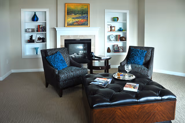 Cozy living room with two patterned armchairs, a tufted leather ottoman, and a fireplace flanked by built-in shelves.