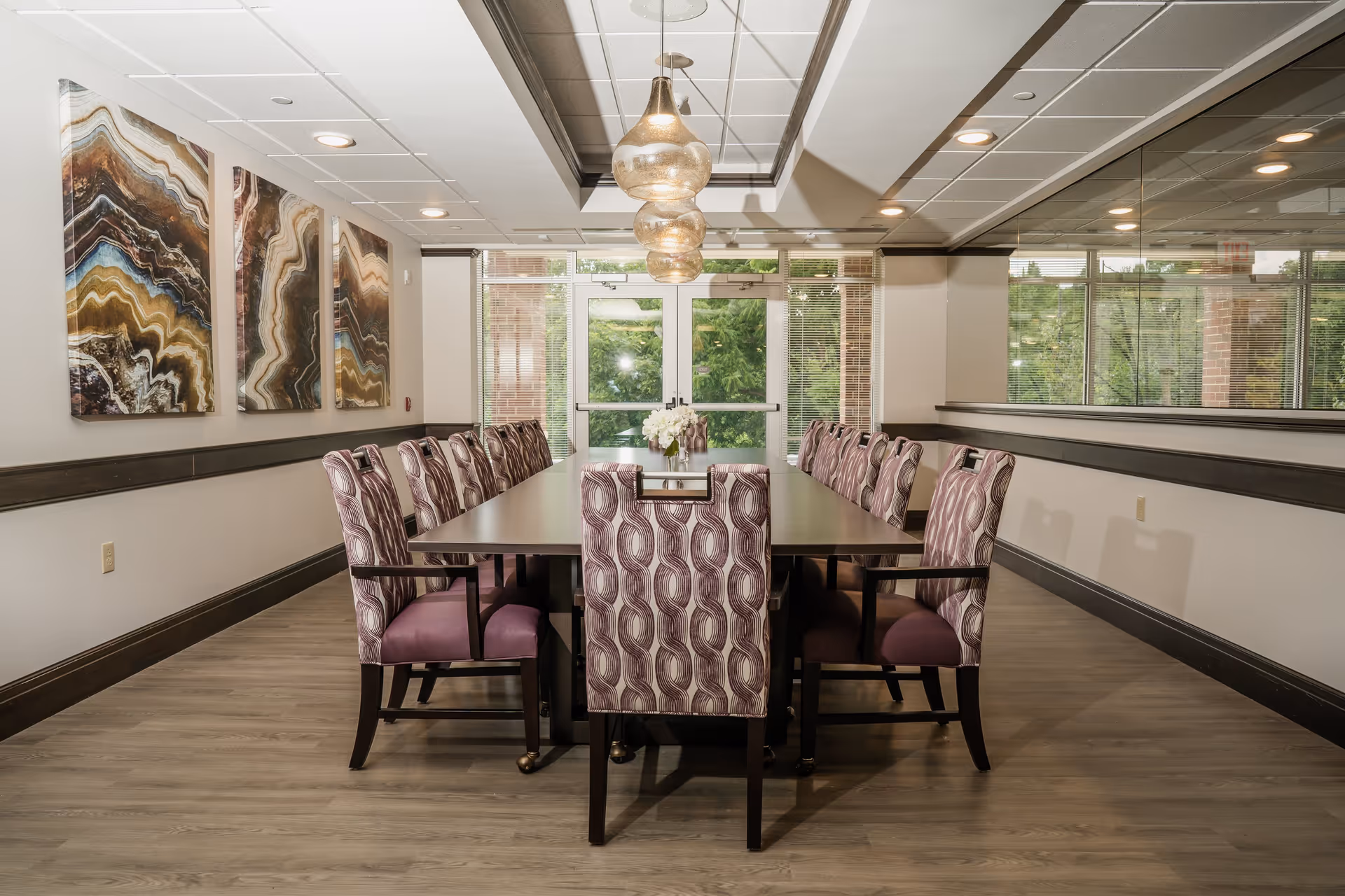 A modern dining room with a long rectangular table surrounded by twelve upholstered chairs with purple patterned fabric. The room features three abstract art pieces on the left wall, large windows and glass doors at the far end letting in natural light, and three pendant lights hanging above the table. The floor is wood, and there is a large mirror on the right wall reflecting the room.
