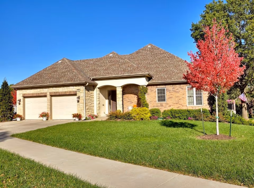 Single-story brick house with a two-car garage, a well-maintained green lawn, and a small tree with red leaves in the front yard under a clear blue sky.