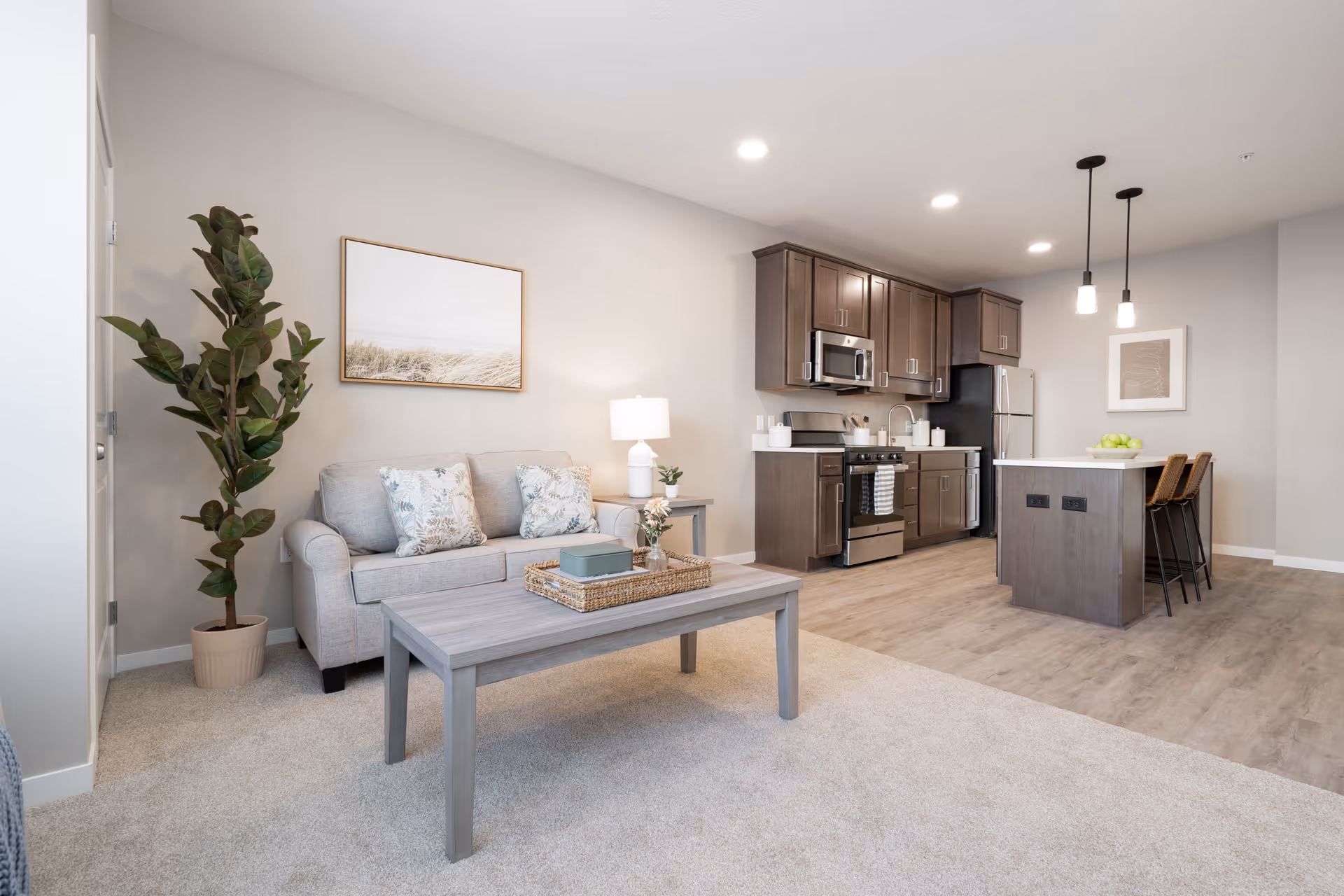 A modern open-concept living area and kitchen in a senior living facility. The living area features a light gray sofa with patterned cushions, a wooden coffee table with decorative items, a tall potted plant, and a framed picture on the wall. The kitchen has dark wood cabinets, stainless steel appliances including a microwave, stove, and refrigerator, and a kitchen island with two bar stools and pendant lights overhead. The flooring transitions from carpet in the living area to wood in the kitchen.