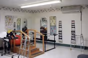 Interior view of a rehabilitation or physical therapy room with parallel bars for walking exercises, a small set of stairs, a walker, exercise balls, and various therapy equipment against the walls.