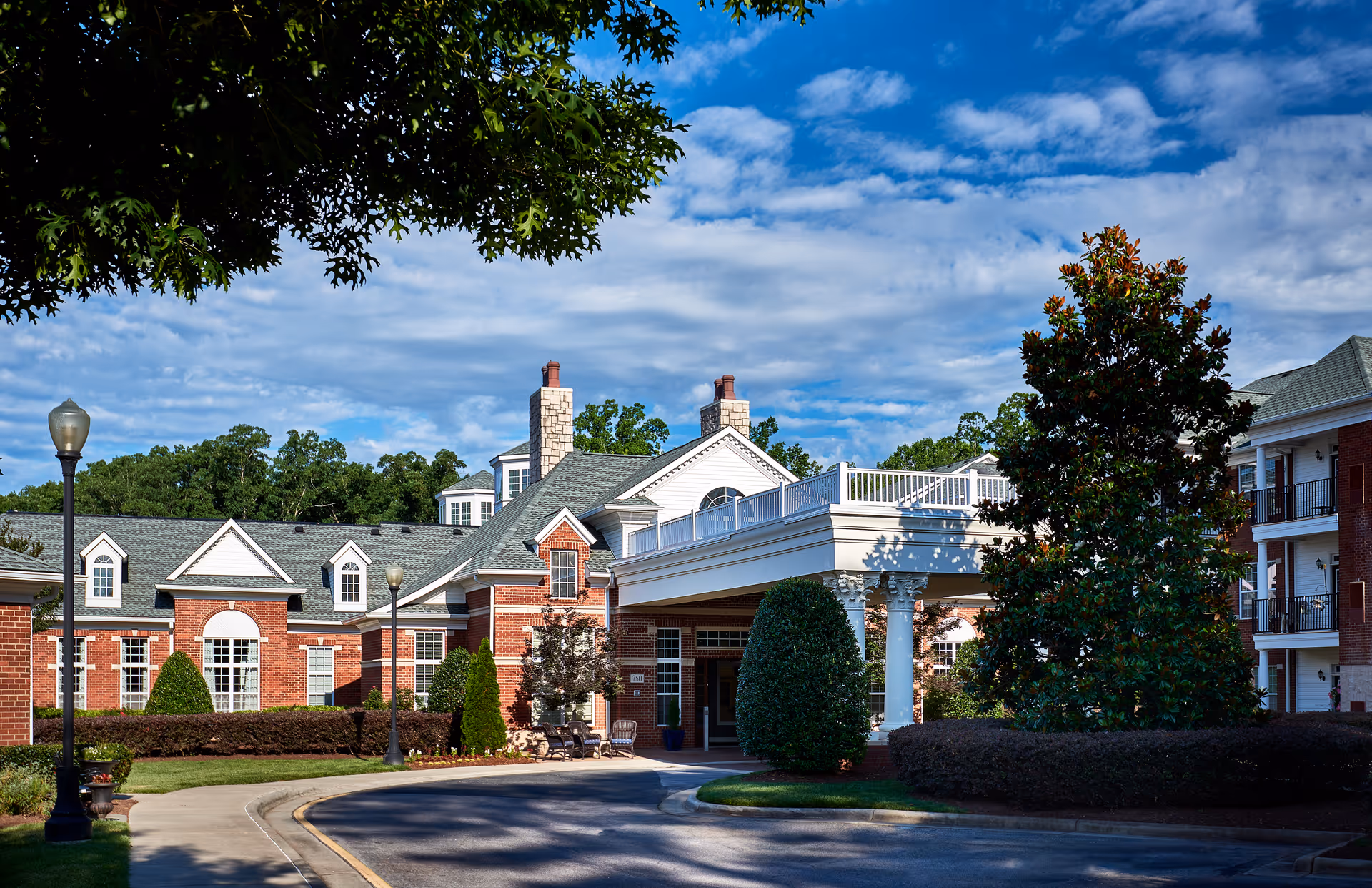 Exterior view of Waltonwood Cary Parkway senior living facility showing a red brick building with white trim, a covered entrance supported by columns, well-maintained landscaping including bushes and trees, and a partly cloudy blue sky.