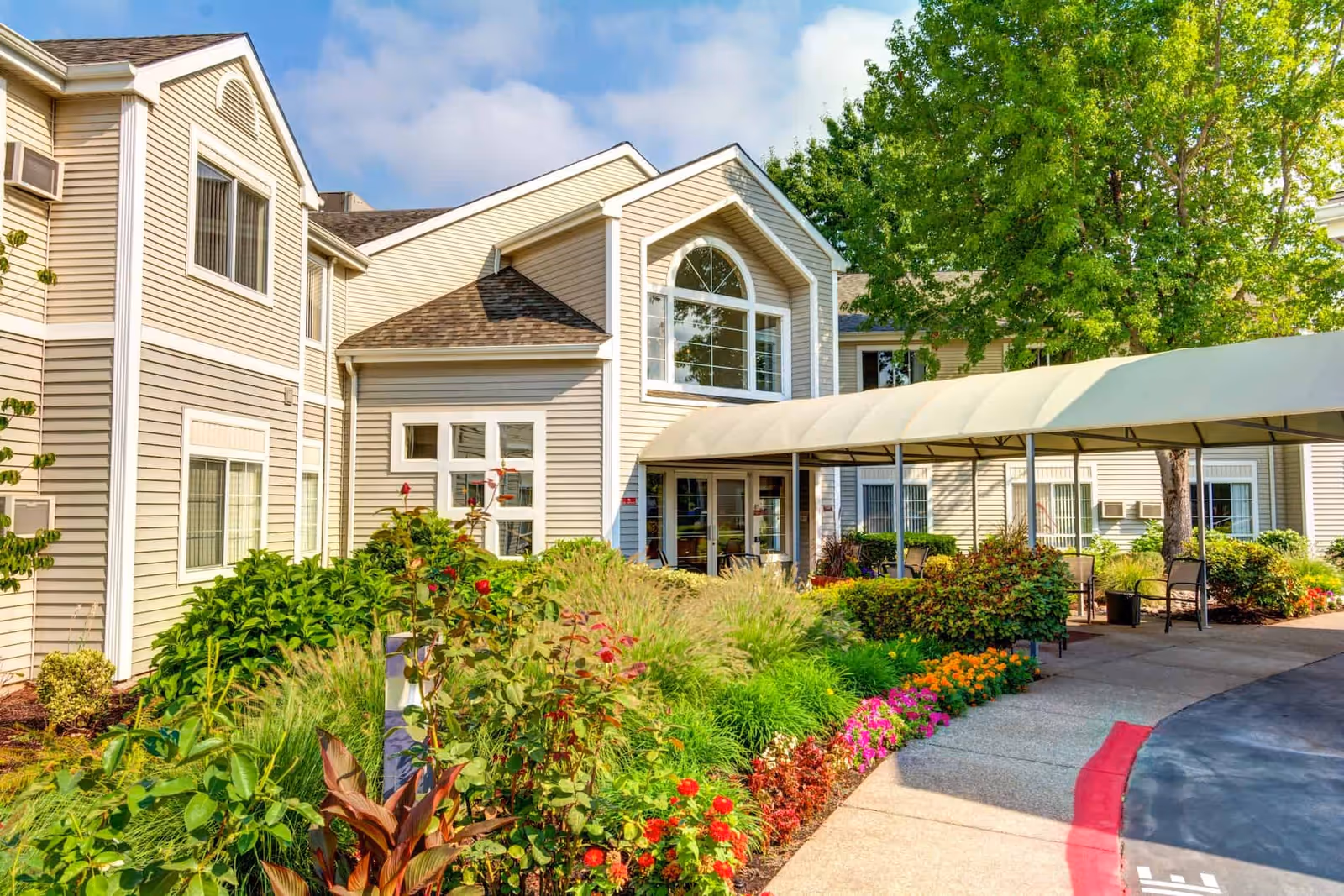 Exterior view of The Oaks at Lebanon senior living facility showing a beige building with large windows, a covered entrance walkway, and a well-maintained garden with colorful flowers and green shrubs under a partly cloudy sky.