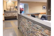 Interior view of a reception or front desk area in a senior living facility with a stone facade on the desk, office chairs, a large floral painting on the wall, and a vase with pink flowers in the background.