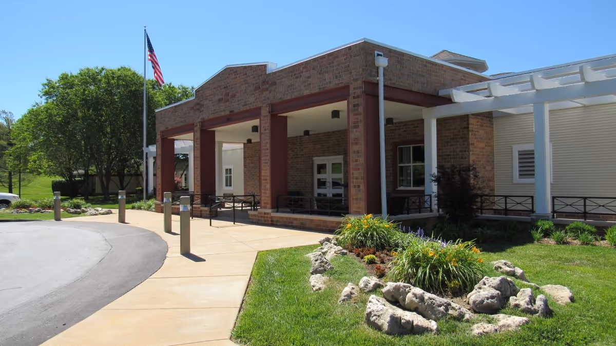 Front entrance of a brick care facility with a covered porte-cochere, landscaped flower beds, an American flag, and a curved driveway.