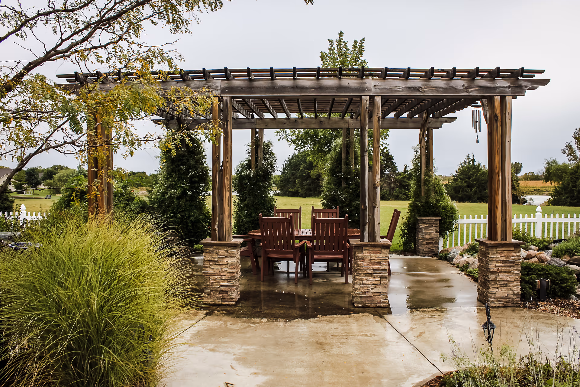 Outdoor wooden pergola with stone pillars covering a patio area with a table and several chairs. Surrounding the pergola are green bushes, trees, and a white picket fence with a grassy field and more trees in the background under an overcast sky.
