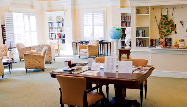 Bright communal sitting and reading room with upholstered chairs, bookshelves, a central table with chairs and informational pamphlets, and a globe.