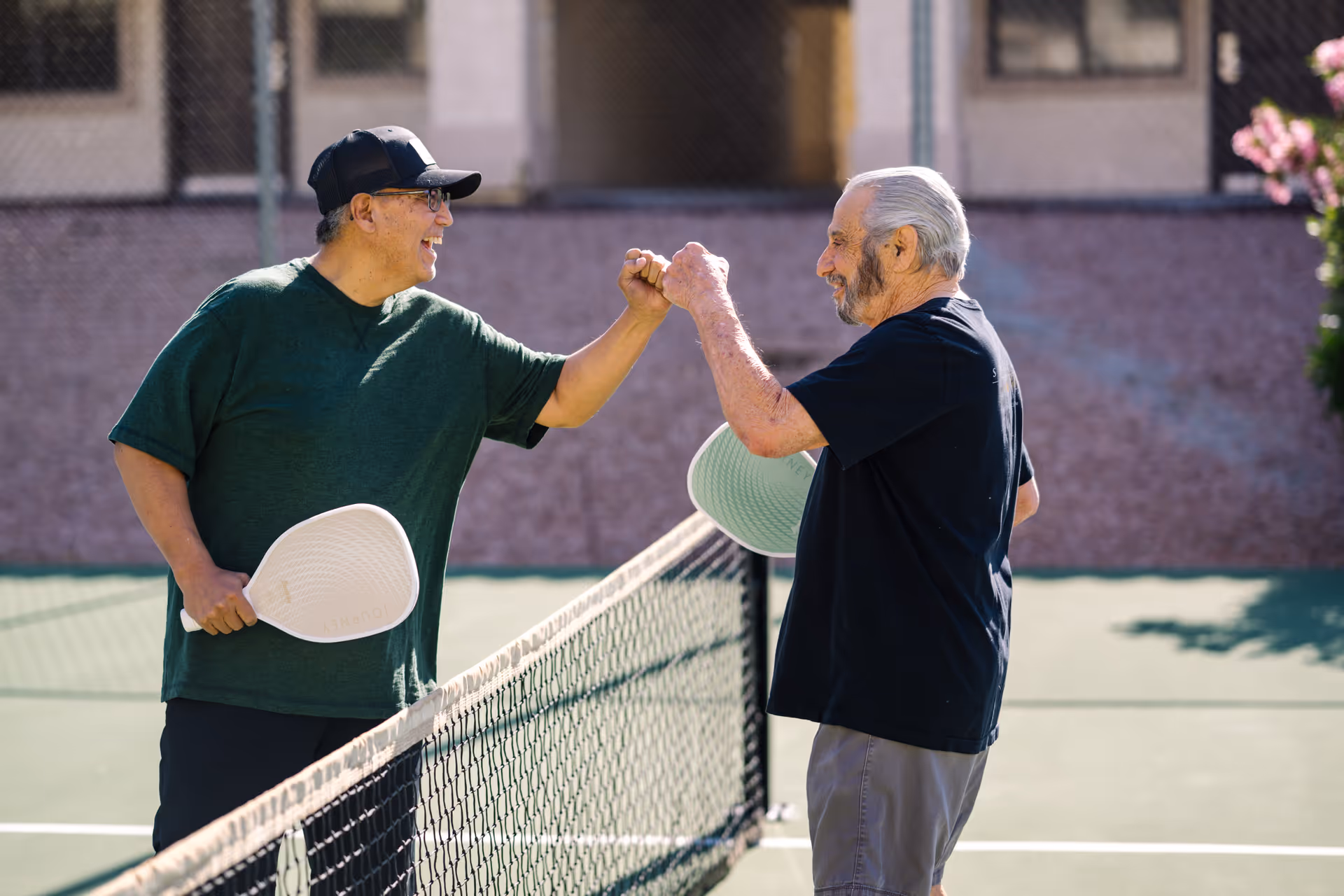 Two older men on an outdoor court fist-bumping over the net while holding paddles.