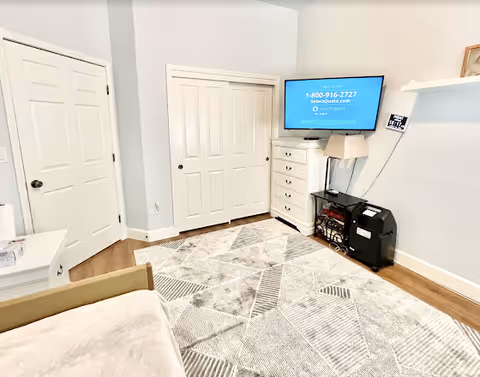 A bright room with light blue walls and wooden flooring, featuring a patterned area rug, a white dresser with a lamp on top, a flat-screen TV mounted on the wall displaying a phone number and website, a small black heater or fireplace, a white shelf with a small framed picture, and two closed white doors including a closet with sliding doors.
