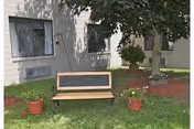 Outdoor area of a facility with a wooden bench on a grassy lawn, two potted plants on either side of the bench, a tree providing shade, and a white building with windows in the background.