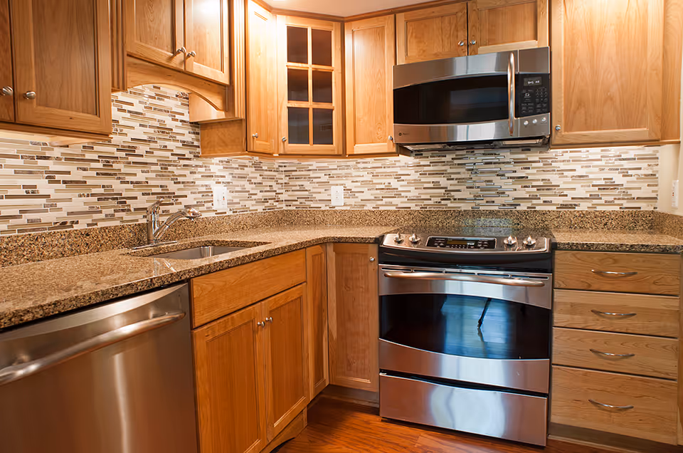 Modern kitchen with wooden cabinets, granite countertops, a stainless steel dishwasher, stove, and microwave. The backsplash features a mosaic tile design in neutral tones.