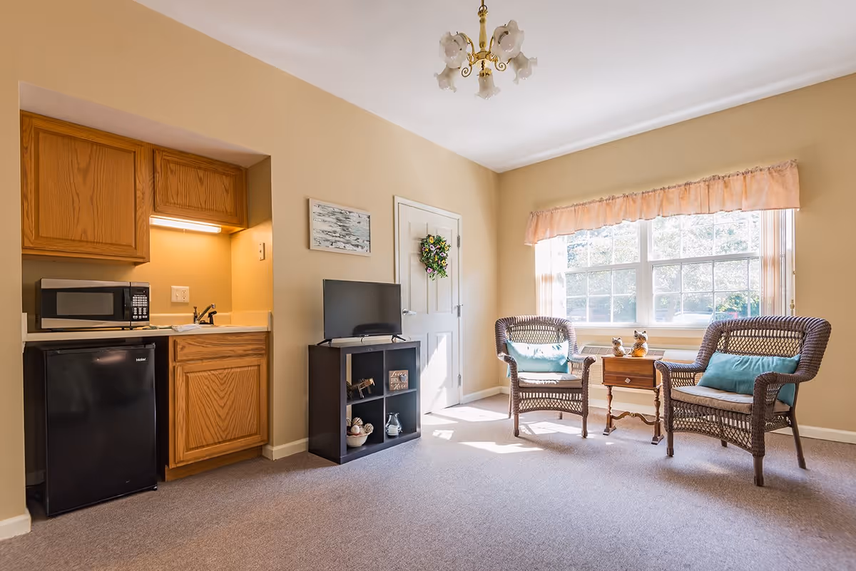 A cozy senior living room with two wicker chairs with blue cushions, a small wooden side table with decorative items, a large window with peach-colored valance curtains letting in natural light, a small black TV stand with a flat-screen TV and decorative items, and a kitchenette area with wooden cabinets, a microwave, a small refrigerator, and a sink.
