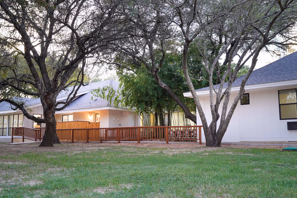 Exterior view of a single-story assisted living facility building with white walls and a gray roof, surrounded by large trees and green grass. There is a wooden railing along a walkway leading to the entrance, and warm light is visible through the windows.