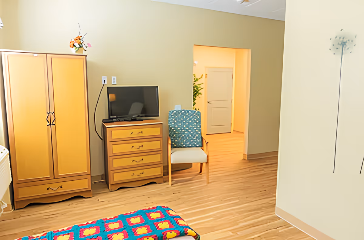 A bedroom in a senior living facility with wooden flooring, a wooden wardrobe, a chest of drawers with a TV on top, a chair with a patterned cushion, and a colorful quilt on the bed. There is a doorway leading to another room with a closed door and some greenery visible.