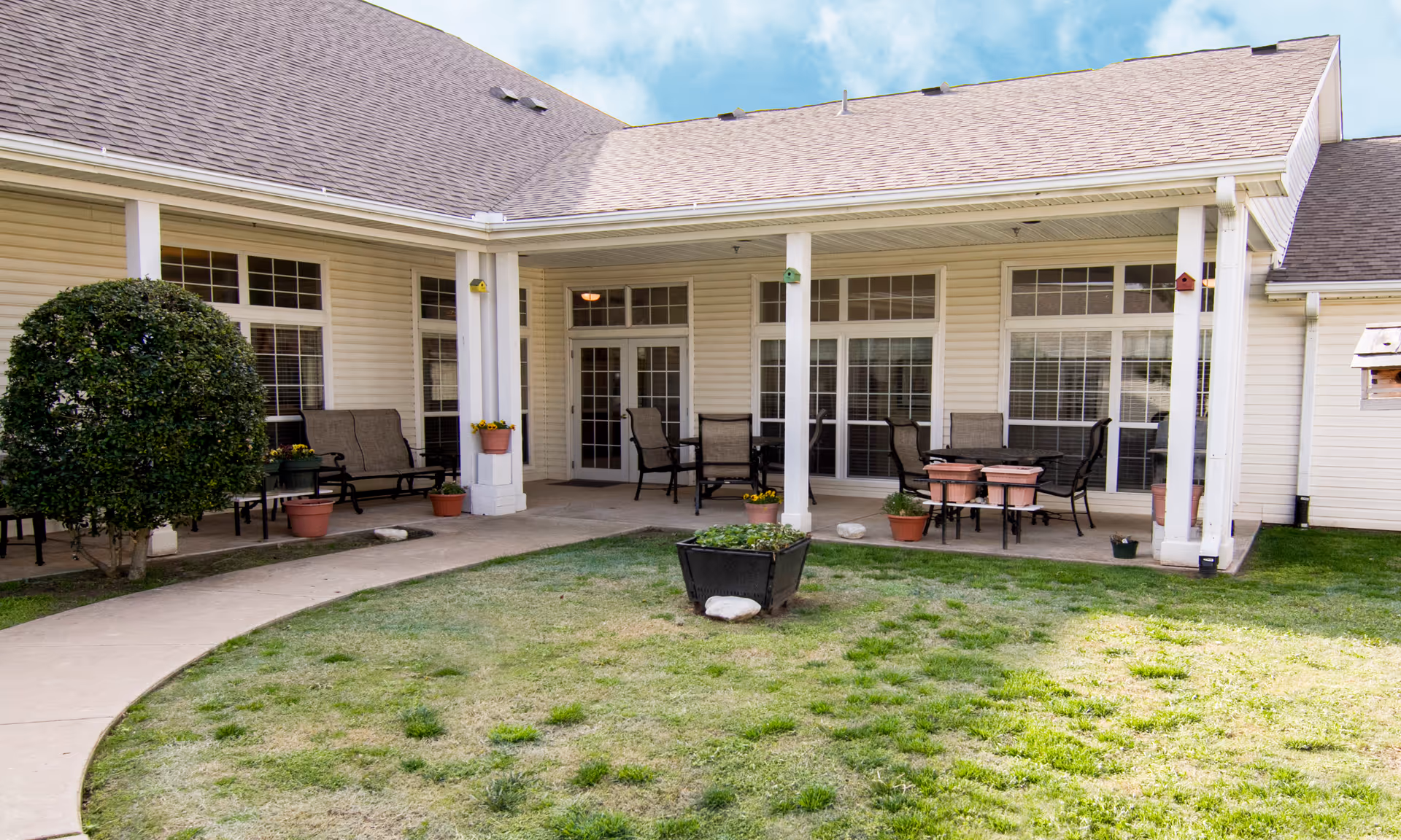 Covered patio with chairs, tables and potted plants outside a single-story assisted living building overlooking a grassy courtyard.