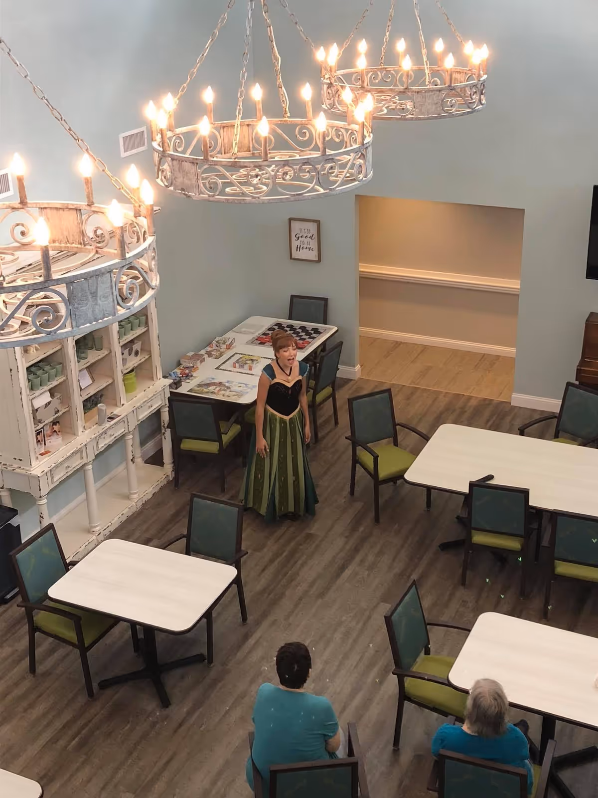 A woman dressed in a green and black costume stands in a room with several tables and chairs. Two people are seated facing her, watching. The room has wooden flooring, light blue walls, and three large decorative chandeliers hanging from the ceiling. There is a white cabinet with cups and games on a table against the wall.