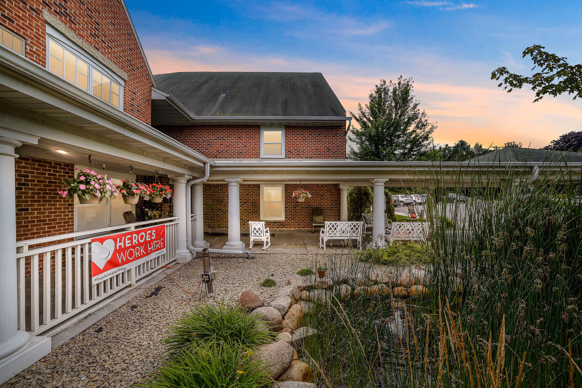 Outdoor courtyard area of a brick building at sunset with white columns and railings. There are hanging flower pots with pink and red flowers, white benches, and a small landscaped garden with rocks and tall grasses. A red banner on the railing reads 'HEROES WORK HERE'.