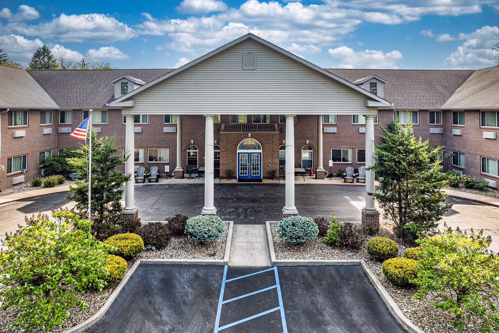Front exterior view of Brentwood at Niles Senior Living facility showing a large covered entrance with white columns, a blue double door, brick building facade, landscaped bushes, and an American flag on a flagpole under a partly cloudy sky.