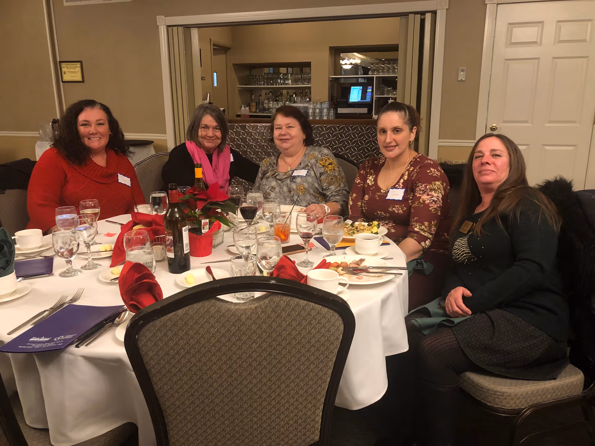 Five women seated around a round banquet table with plates, glasses, and wine at an indoor dining event.