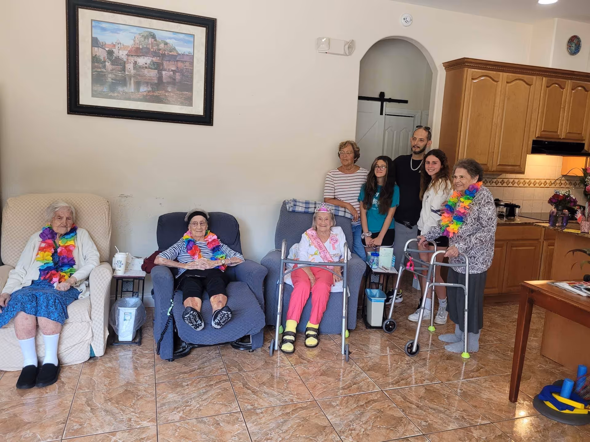 A group of elderly women and younger adults gathered in a living room area. Three elderly women are seated in armchairs, two wearing colorful leis and one wearing a tiara and sash. Two elderly women are standing, one using a walker and also wearing a colorful lei. Three younger adults stand behind the seated women. The room has tiled floors, wooden kitchen cabinets, and a framed painting on the wall.