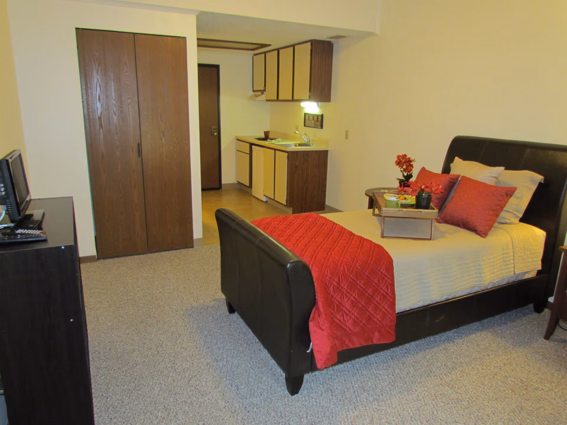 A studio apartment room with a bed featuring a beige bedspread and red pillows and blanket. A small wooden tray with flowers and a cup is on the bed. There is a dark wooden dresser with a TV and remote on it. In the background, there is a kitchenette with cabinets, a sink, and a small refrigerator. A wooden closet door and an entrance door are also visible.