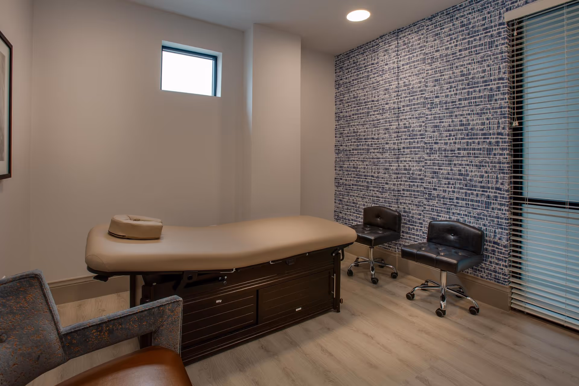 A small treatment room with a beige massage or examination table in the center, two black swivel chairs against a blue patterned accent wall, a window with blinds on the right, and a small high window on the left wall.