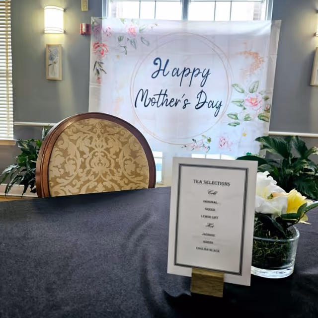 A decorated table with a black tablecloth, a floral-patterned chair, a small sign listing tea selections, and a glass vase with white flowers. In the background, a banner reads 'Happy Mother's Day' with floral designs, set against a window with natural light and plants on either side.