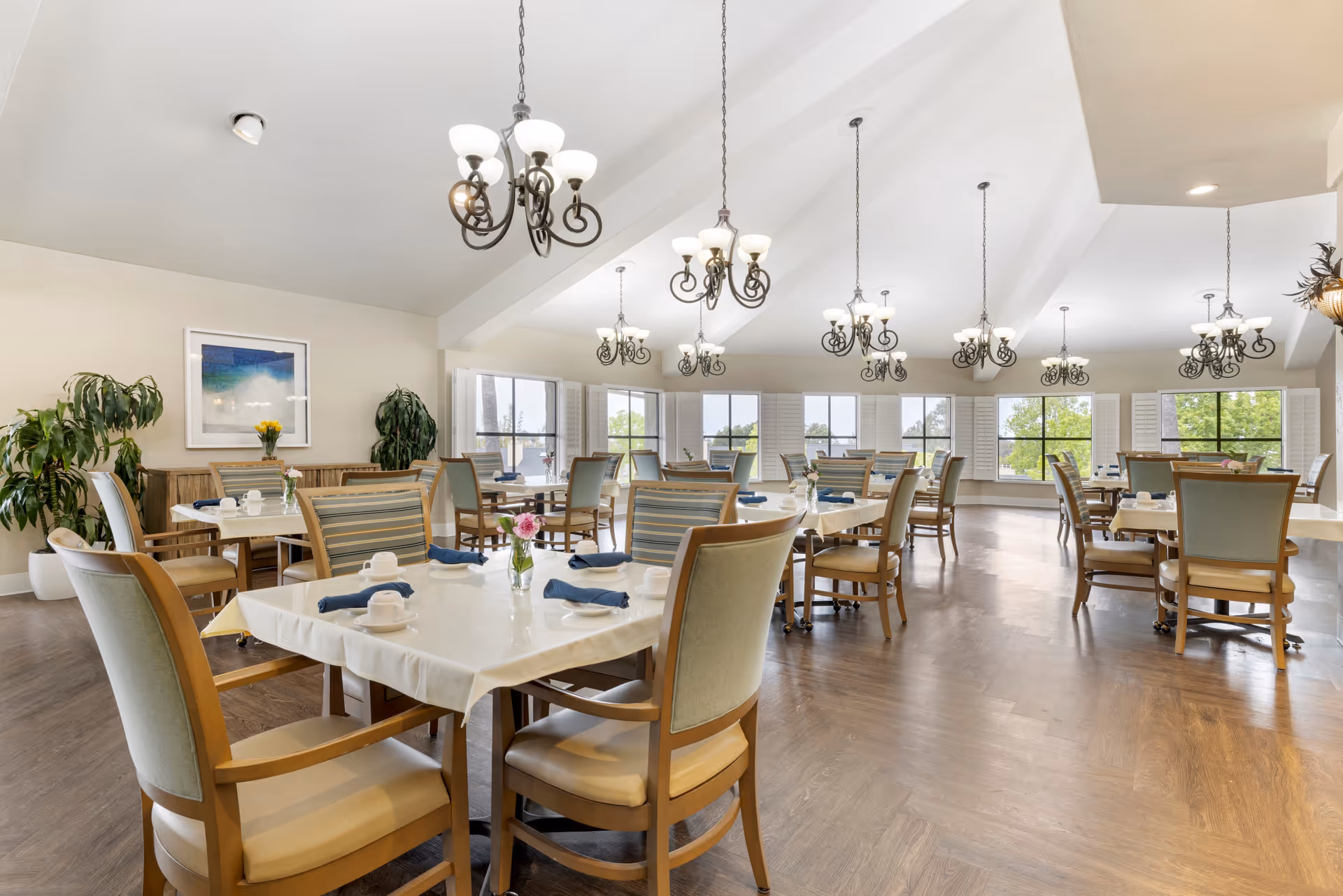 A bright and spacious dining room with multiple tables covered in white tablecloths, each set with cups, plates, and blue napkins. The room features wooden chairs with cushioned seats and backs, large windows with white shutters letting in natural light, several hanging chandeliers with multiple lights, and potted plants along the walls.