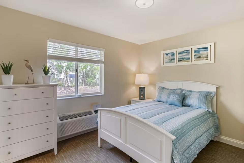 Bright bedroom with white furniture, blue bedding, and a large window overlooking greenery.