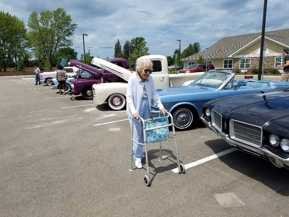 An elderly woman using a walker is standing in a parking lot with several classic cars and trucks lined up with their hoods open. There are a few other people in the background near the vehicles, and a building with a sloped roof is visible behind the parking lot under a partly cloudy sky.
