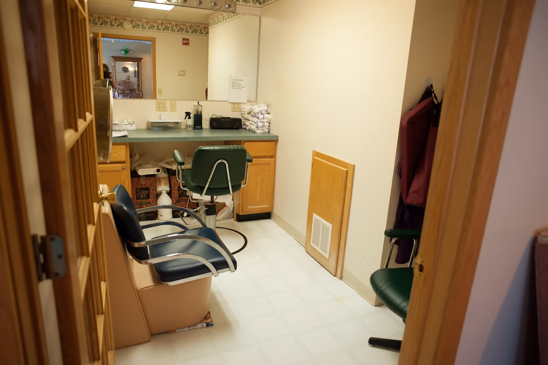 A small salon or grooming room with two salon chairs, a countertop with various supplies including towels, a spray bottle, and a radio. There is a large mirror on the wall above the countertop, and the room has light-colored walls and flooring with wooden cabinetry.