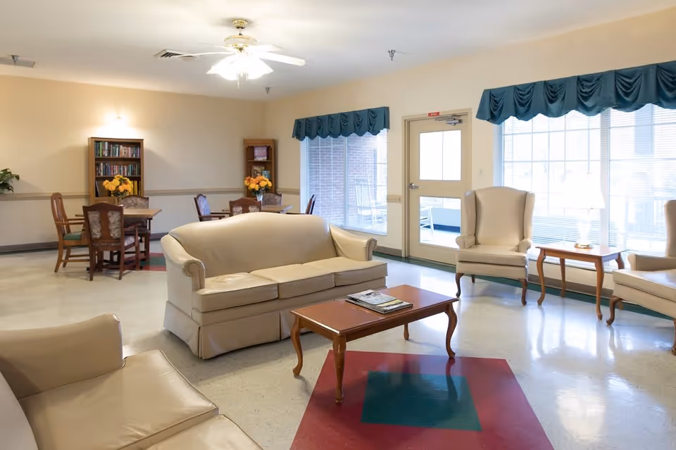 A bright and spacious living room area in a senior living facility with cream-colored leather sofas and armchairs arranged around a wooden coffee table. There are two small wooden tables with lamps near large windows covered with teal valances. In the background, there are two small dining tables with chairs and a bookshelf filled with books and decorative items. The floor has a pattern with red and green squares, and a ceiling fan with lights is mounted above.