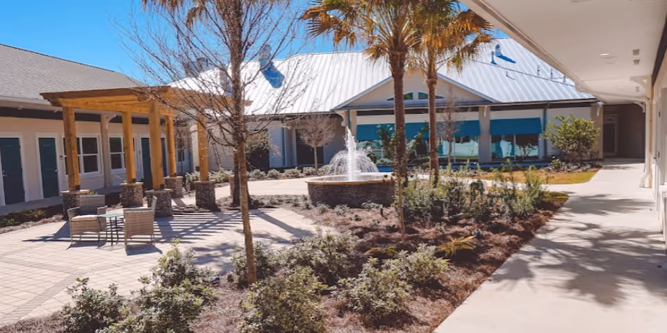 Outdoor courtyard area of A Banyan Residence Assisted Living Resort featuring a paved patio with chairs and tables, a wooden pergola, landscaped garden beds with shrubs and palm trees, and a central water fountain. The buildings surrounding the courtyard have white walls and teal window awnings under a metal roof.
