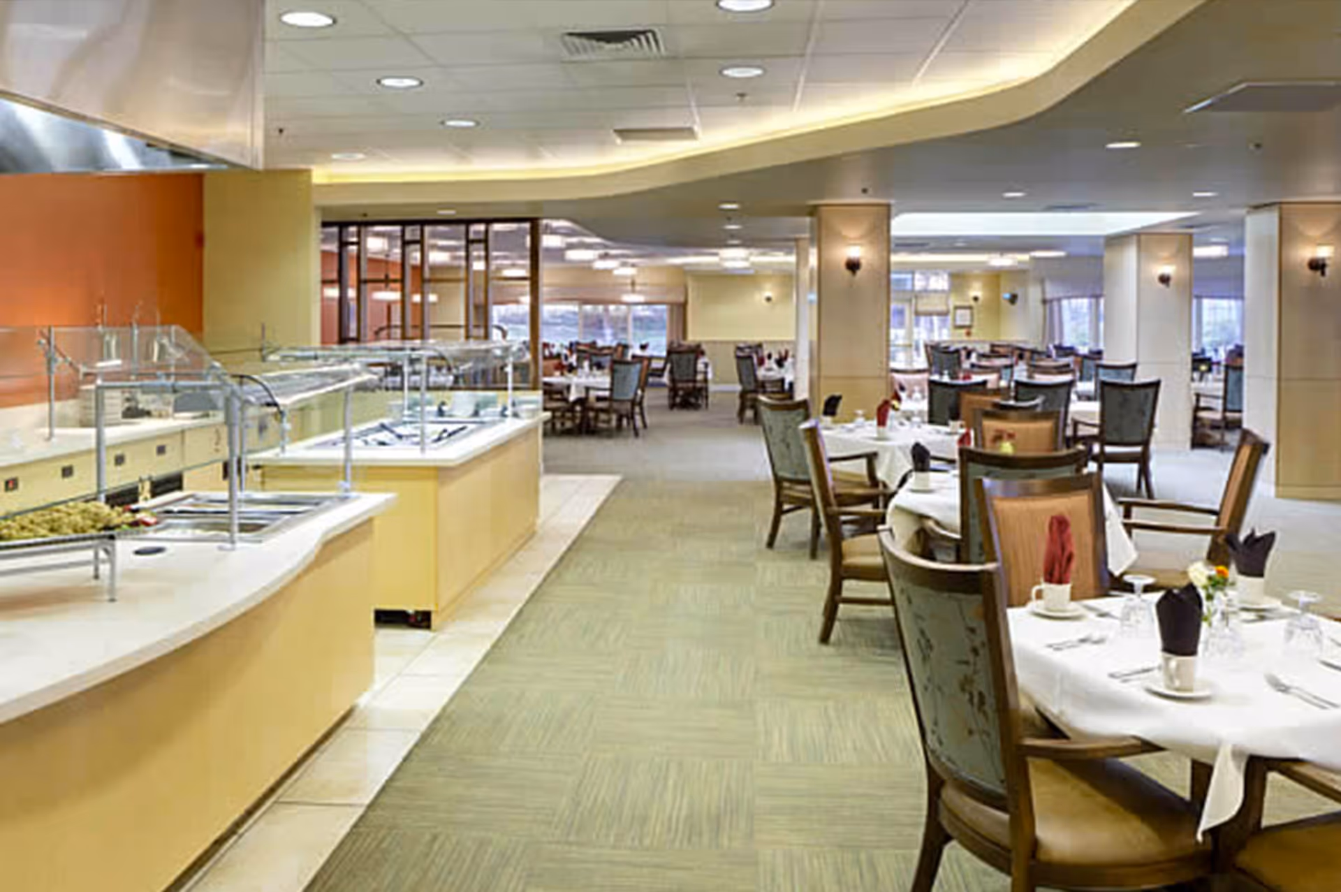 Interior view of a spacious dining room with multiple round tables set with white tablecloths, napkins, cups, and glassware. To the left, there is a buffet serving area with food trays and sneeze guards. The room has carpeted flooring, ceiling lights, and large windows in the background.