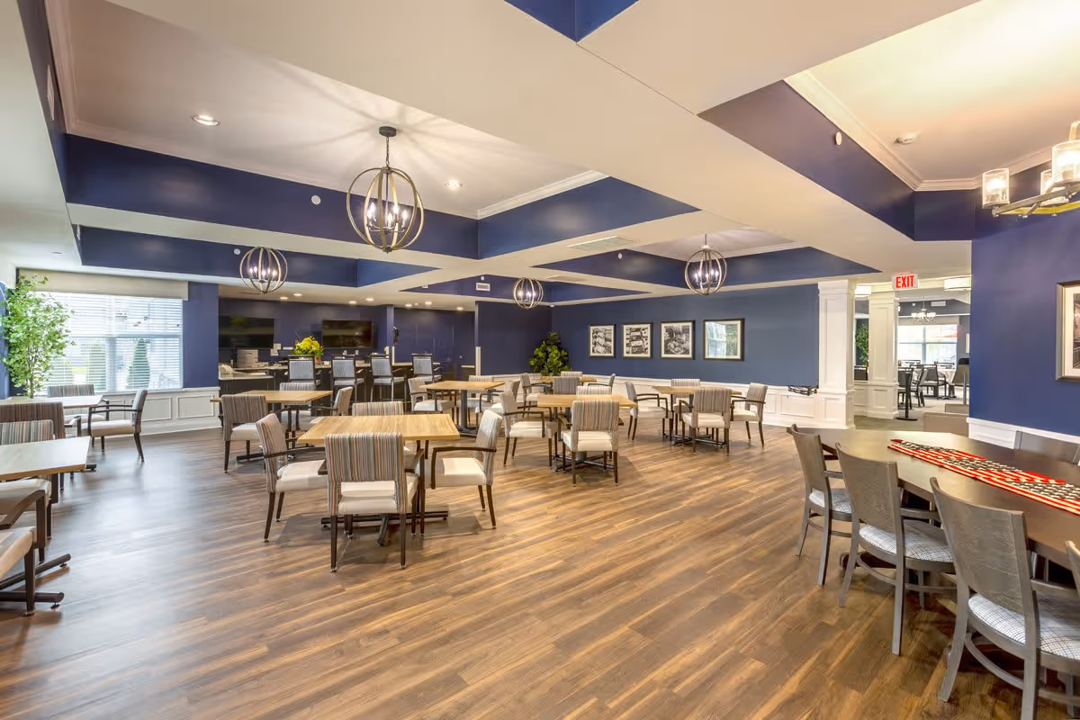 A spacious dining room with multiple wooden tables and cushioned chairs arranged neatly on a wood floor. The walls are painted blue with white wainscoting, and there are modern spherical pendant lights hanging from the ceiling. Large windows allow natural light to enter, and framed black and white pictures decorate one wall. There is an exit sign above a doorway leading to another room.