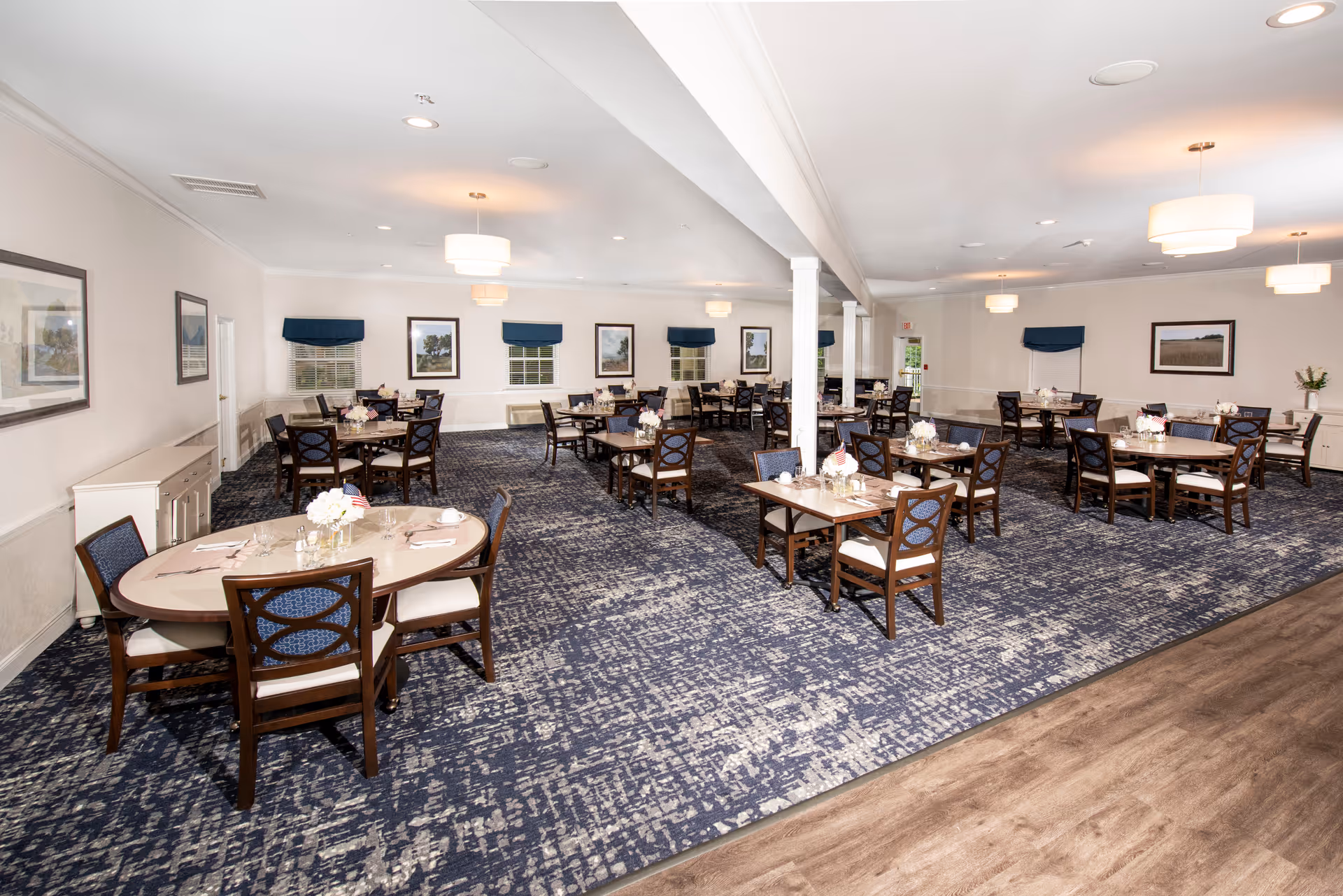 A spacious dining room in a senior living facility with multiple tables and chairs arranged neatly. Each table is set with glassware, napkins, and small floral centerpieces. The room features blue patterned carpet, white walls adorned with framed landscape paintings, and several windows with blue valances. Ceiling lights provide warm illumination, and there is a wooden floor area visible in the foreground.