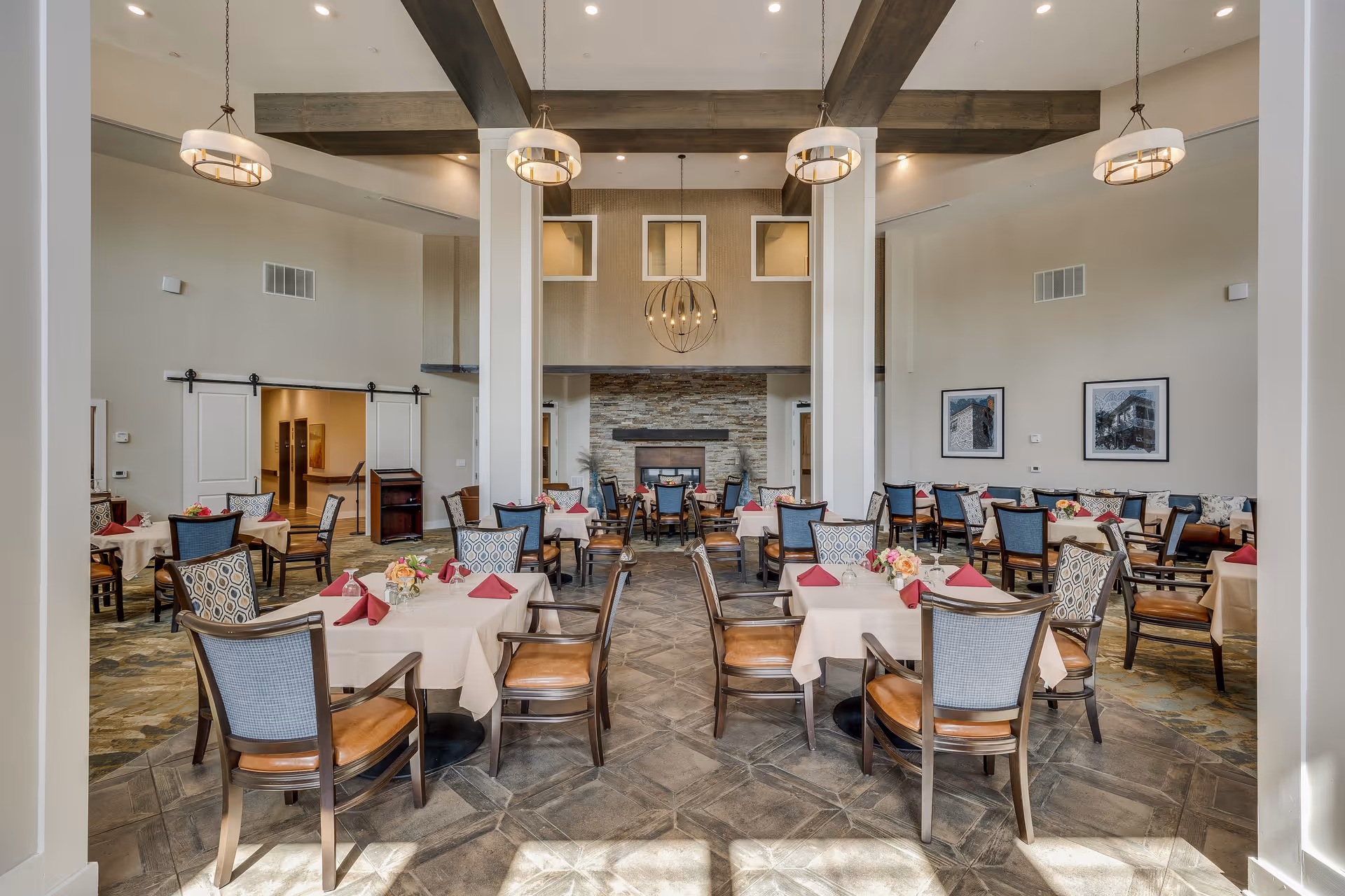 A spacious dining room with multiple tables covered in white tablecloths and set with red napkins and floral centerpieces. The room features high ceilings with exposed wooden beams, modern hanging light fixtures, and a stone fireplace at the far end. Chairs with wooden frames and cushioned seats surround the tables. The walls are decorated with framed pictures.