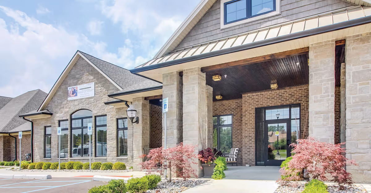 Front entrance of a stone and brick assisted living building with columns, large windows, and landscaped shrubs.
