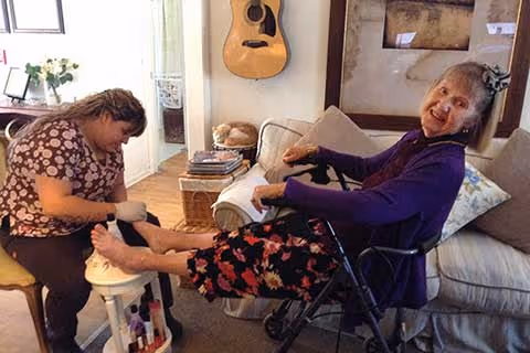 An elderly woman sitting on a couch with a walker in front of her, smiling and receiving a foot massage or pedicure from a caregiver seated on a stool. The room has a guitar hanging on the wall and a small table with books and decorative items.
