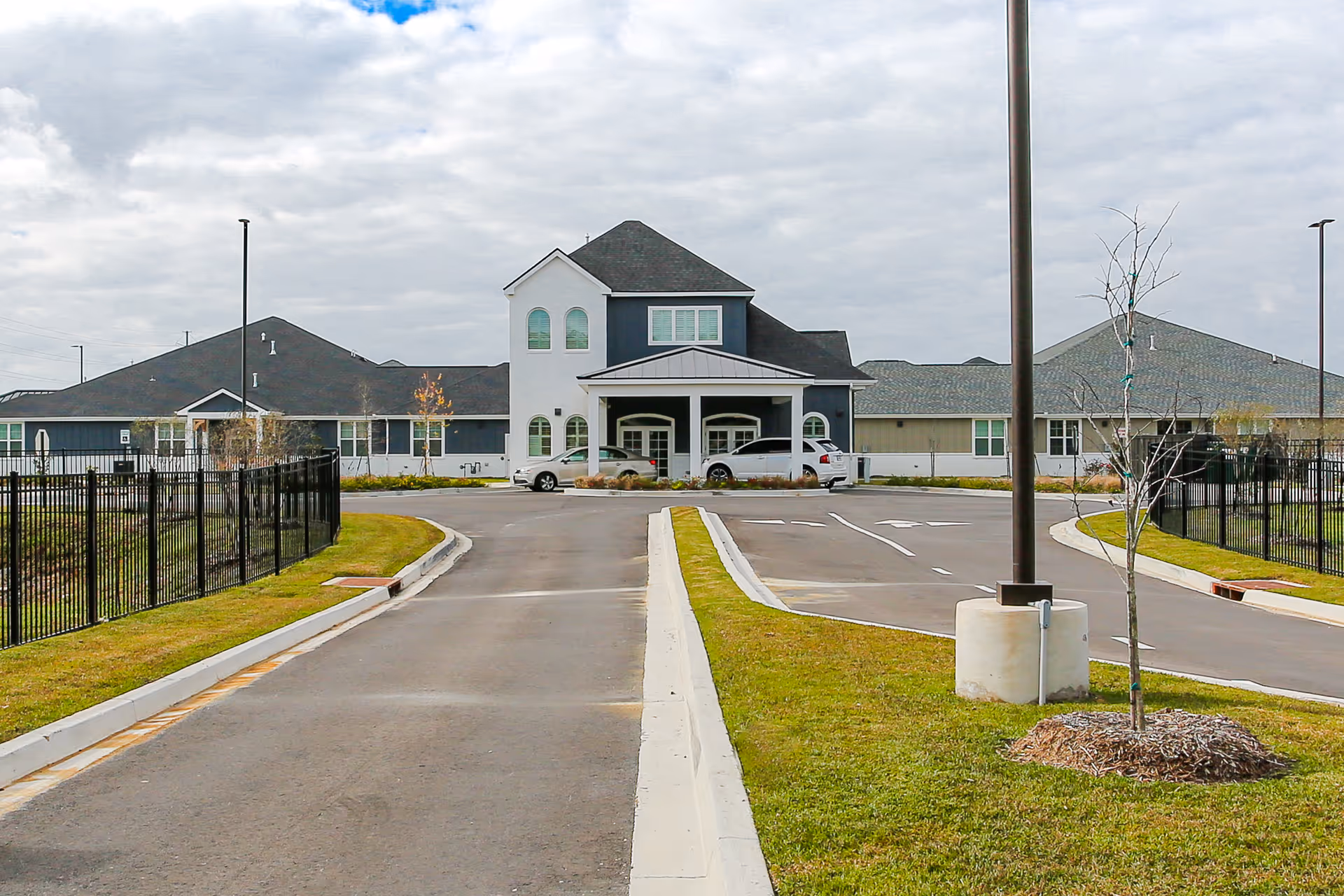 Front exterior view of a senior living facility named Sage Oak of Lake Charles, showing a large building with a covered entrance, multiple windows, and a driveway with parked cars. The sky is cloudy and there is a grassy area with a small tree and a lamp post in the foreground.