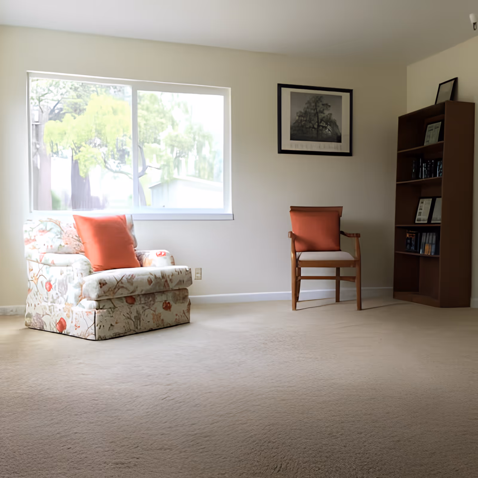 A bright room with a large window showing green trees outside. Inside, there is a floral patterned armchair with an orange cushion, a wooden chair with an orange cushion, a wooden bookshelf with books and framed pictures, and a framed black and white picture hanging on the wall.