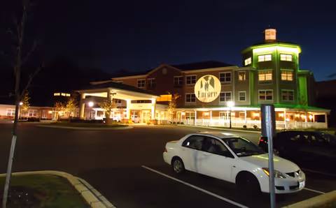 Nighttime exterior view of the Lorien Encore senior living facility with the building illuminated by lights, including a green-lit tower section and a large circular sign displaying the facility's name. A white car is parked in the foreground in an empty parking lot.