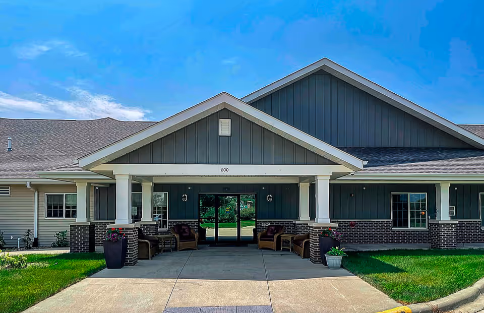 Front exterior view of a single-story senior living facility building with a covered entrance supported by white columns. The building has gray siding with brick accents and a peaked roof. There are chairs and potted plants near the entrance, and a clear blue sky above.