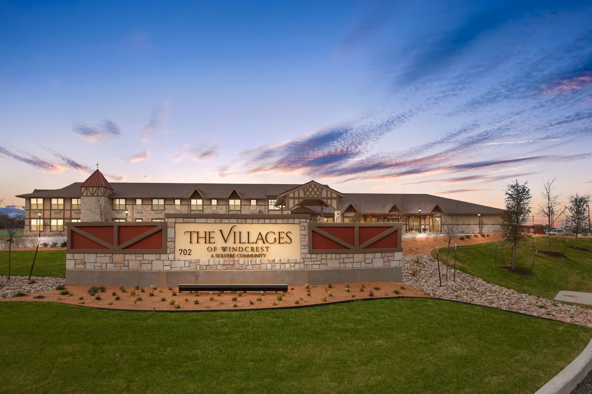 Exterior view of The Villages of Windcrest building at dusk with a large stone sign in front displaying the facility name and address, surrounded by green grass and landscaping under a partly cloudy sky.