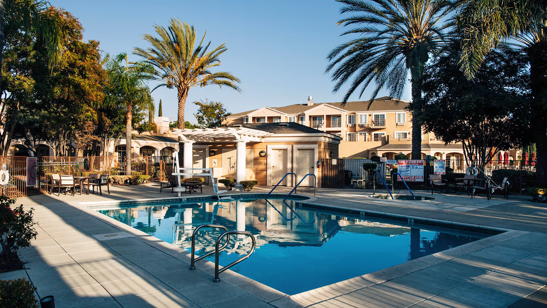 Outdoor swimming pool area at Atria Park of Vintage Hills with clear blue water, surrounded by palm trees, patio chairs, tables, and a small building with a pergola. The background shows a multi-story residential building under a clear blue sky.