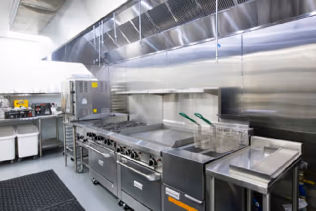 A commercial kitchen with stainless steel appliances including a stove, oven, griddle, deep fryer, and ventilation hood. There are also sinks and preparation areas visible, with a black anti-fatigue mat on the floor.
