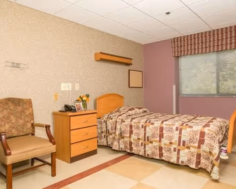 A single bed with a patterned quilt in a small room. Next to the bed is a wooden nightstand with a telephone, a framed photo, and a small vase of flowers. There is a cushioned armchair with wooden arms to the left of the nightstand. The walls are painted beige and purple, and a window with blinds and a valance is on the right side of the room.
