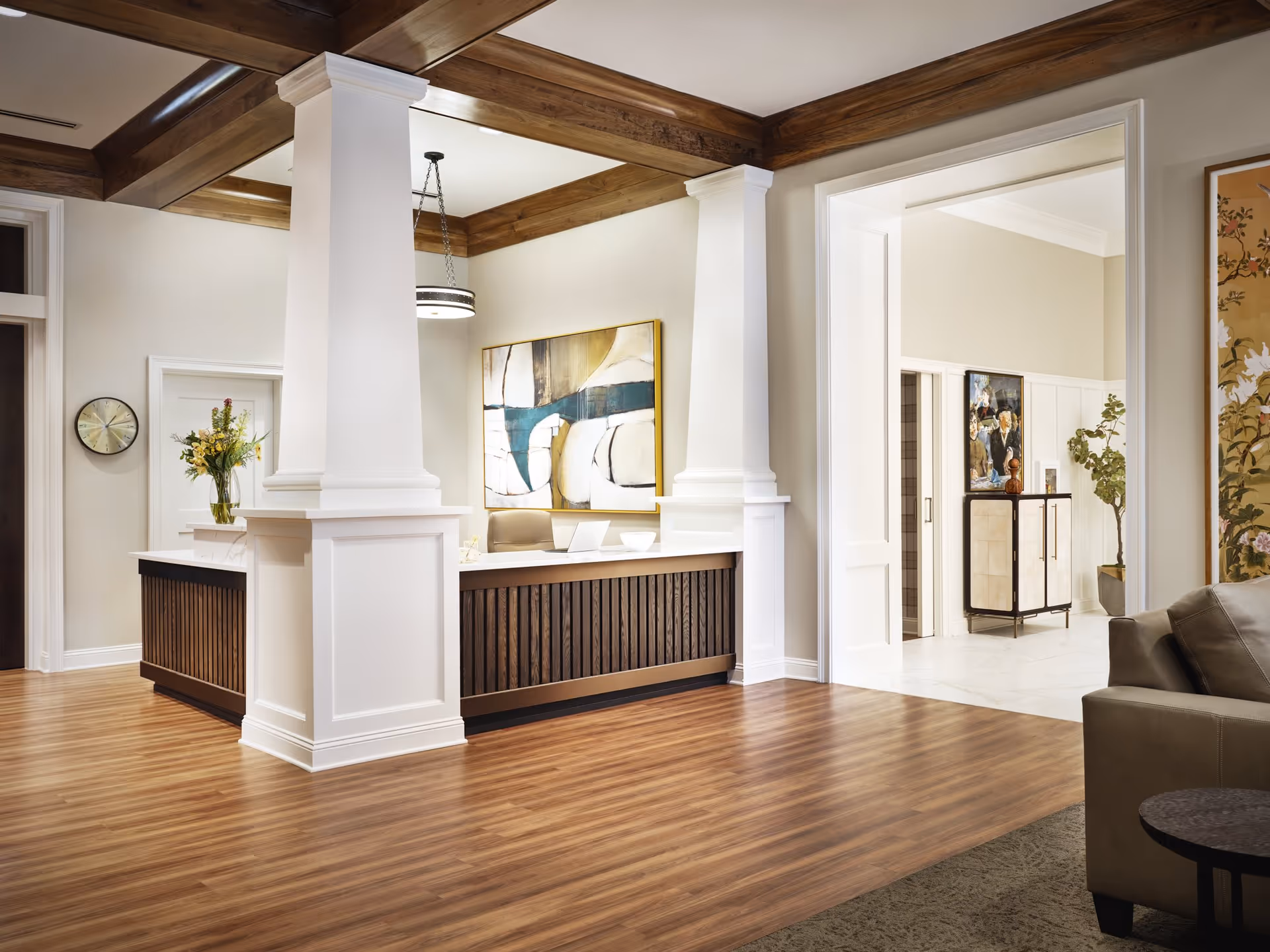 A spacious and elegant senior living facility reception area with wooden flooring, white pillars, and a wooden reception desk. The walls are light-colored with a large abstract painting behind the desk. There is a vase with flowers on the desk, a clock on the wall, and a glimpse of a hallway with artwork and plants in the background. A portion of a leather sofa and a small round table are visible in the foreground.